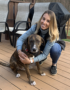 A woman wearing a long-sleeve blue shirt and jeans, has long blonde hair. She is squatting down on a wooden deck with her arms around a medium size dog.