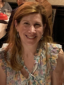A woman with long reddish brown hair smiles. She is wearing a flowered blouse.