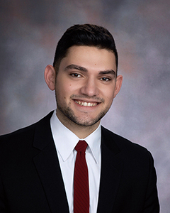 A man with short dark hair, wearing a dark sit, white shirt and red tie, smiles in a formal portrait.