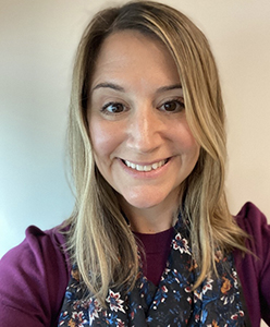 A woman with  longer blonde hair smiles. She is wearing a purple shirt.