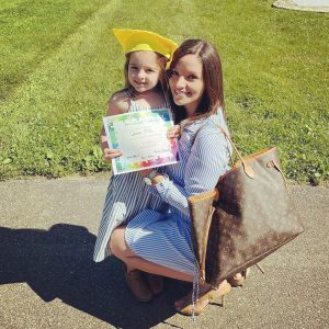 A woman with long brown hair holding a large purse squats down as a little girl earing a yellow graduation cap and holding a certificate sits on her lap. They are both smiling.