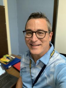 A man in a blue shirt and round glasses smiles. 