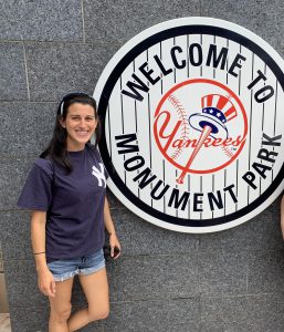 A woman in a navy blue t-shirt with the Yankees NY emblem on it stands in front of a pinstripe sign that says Welcome to Monument Park with the Yankees red white and blue emblem in it.