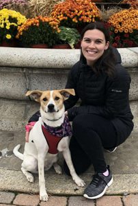 A woman wearing a dark jacket and pants squats down in front of a low wall with her dog, a brown and white little dog with a red harness on.