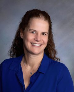 A woman with shoulder-length curly dark hair smiles. She is wearing a dark purple blouse and necklace.