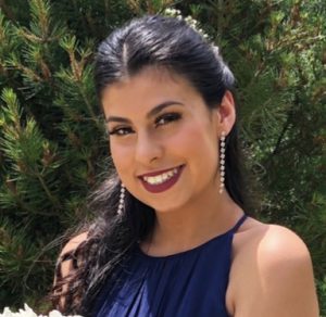 A young woman with long dark hair swept to one side smiles and wears a dark blue halter dress.