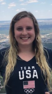 A young woman with long blonde hair. She is smiling and wearing a Team USA blue shirt with the flag on it.