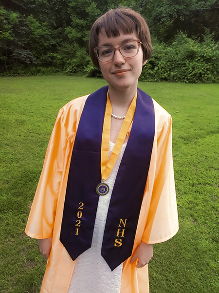 A young woman with short dark hair and glasses wearing a gold graduation gown and a blue wrap around her shoulders that says 2021 NHS