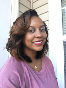 A woman with shoulder'length brown hair smiles. She is wearing a light purple shirt and a gold necklance.