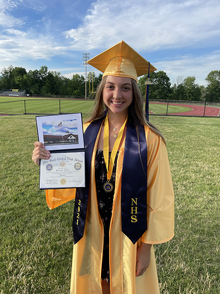 A young woman with longer blonde hair wearing a gold graduation cap and gown. She is holding a diploma and she is smiling. She is wearing a blue wrap around her shoulders that says 2021 NHS