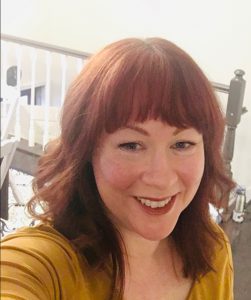 A woman with shoulder-length red hair, wearing a gold shirt. She is smiling.