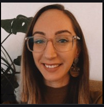 A woman with long brown hair smiles. She is wearing large style glasses and a dark shirt.