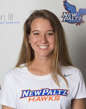 A young woman smiles broadly. She has long blonde hair and is wearing a white t-shirt that says New Paltz Hawks.