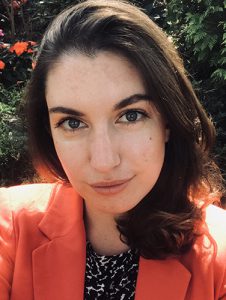 A woman with long dark hair is wearing an orange blazer. She is smiling.