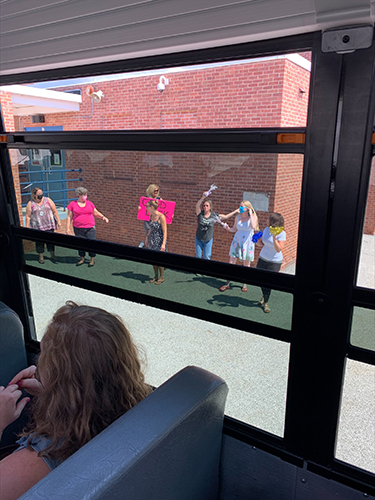 Several people stand outside of a building waving and holding signs that say welcome. The picture is taken through the window of a bus.