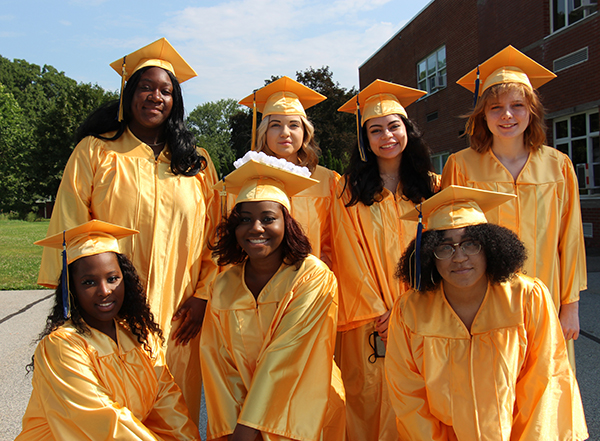 Seven young women, all dressed in gold caps and gowns stand together. There are four in the back and three in the front. All are smiling.