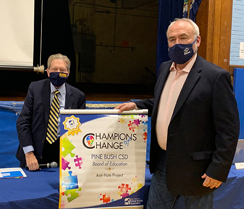 A man wearing a dark suit, blue shirt and blue and gold striped tie accepts a banner that says Champions for Change on it from a man wearing a dark jacket and light colored shirt. Both are wearing dark masks.