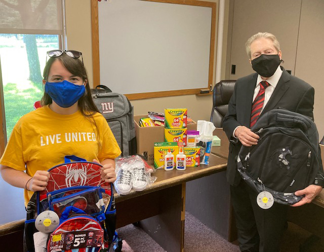 A woman on the left wearing a yellow shirt and blue mask holds a back pack while a man on the right wearing a dark suit, red tie, black mask holds another back pack. Between them is a table filled with schools supplies including crayons, paper, markers.