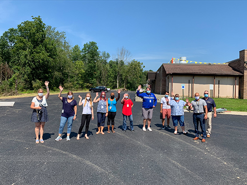 A group of 10 people standing outside on blacktop, waving and smiling.