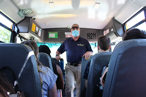 A man wearing a blue shirt and blue mask stands inside of a bus facing the people who are sitting down.