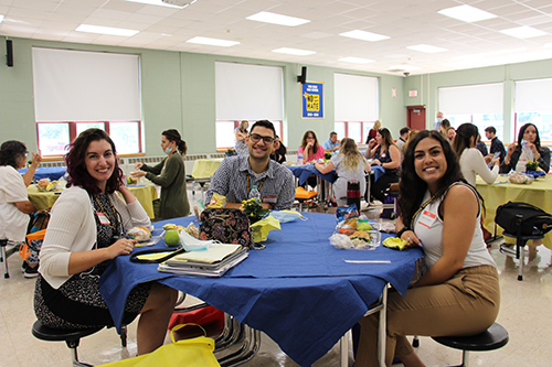 A group of three people, two women and a man, sit at a round table with a blue tablecloth. There are other tables with people around them. They have food in front of them.