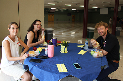 Two women and one man sit at a round table with a blue tablecloth. There is food on the table and water bottles.