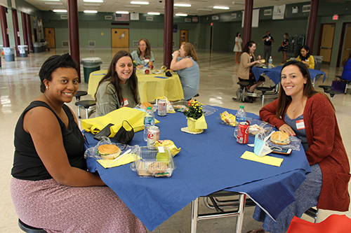 Three women sit at a round table eating lunch. There is a blue table cloth on the table along with food and water bottles. The women are smiling and there are other tables in the background.