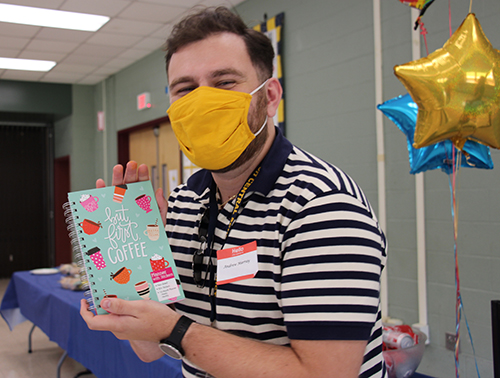 A man in a striped shirt with a yellow face mask holds up a book.