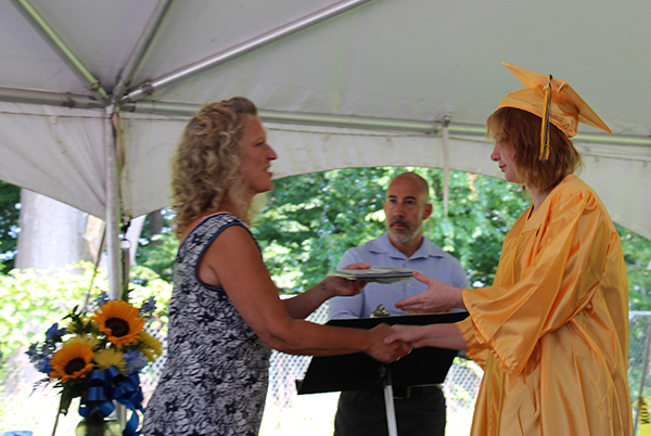 A woman with shoulder-length blonde hair hands a diploma to a young woman in a gold cap and gown. They are shaking hands. A man stands behind them watching.