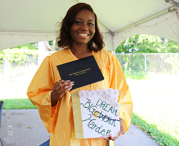 A young woman with long dark wavy hair smiles broadly holding a diploma and decorated graduation cap. The cap says dream big work harder. She is wearing a gold graduation gown.