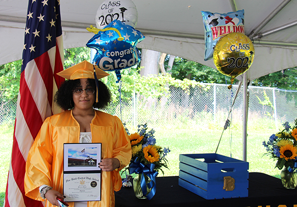 A young woman in a gold cap and gown stands holding her diploma. She has chin length dark hair and is standing in front of a table with flowers and graduation balloons.