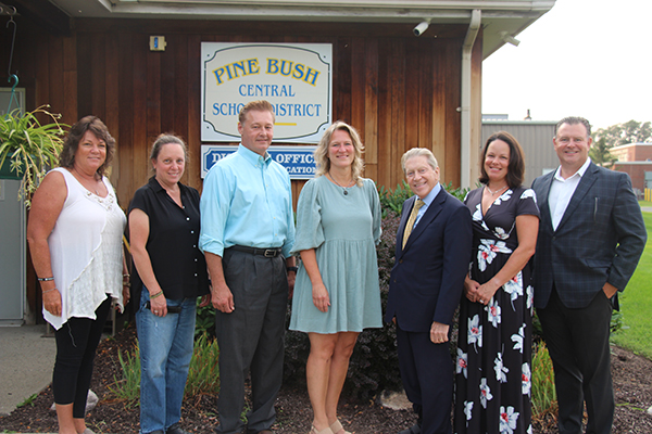 In the background is a wood sided building with the sign Pine Bush Central School District in the center. In front of the building are seven adults, four women and three men in a line, each turned sideways just a bit.