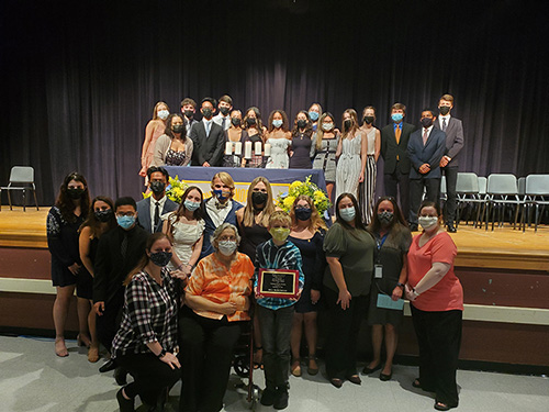 16 high school students dressed nicely in dresses and suits stand on the stage in an auditorium. On the floor below, there are 15 more people. All are wearing masks.