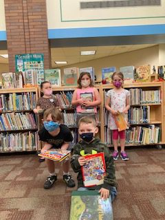 With filled bookshelves behind them, five elementary students stnad holding books up for al to see. They all are wearing masks.