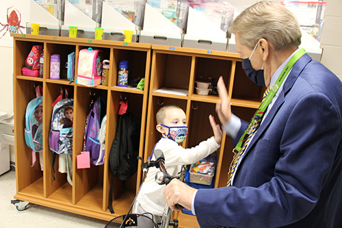 A pre-K boy gives an "air" high five to a man in a blue suit. The boy has a cream colored long-sleeve t shirt on with a mask. Behind him are little cubbies with backpacks and lunch bags.