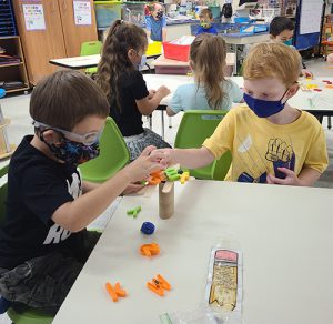 Two boys, one with reddish hair and wearing a blue mask and yellow shirt, and another with a black shirt, black print mask and glasses, put magnetic letters on a cardboard tube fitted with popsicle sticks as branches. There are girls in the background sitting at another table doing the same.