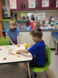 TWo kindergarten boys, one wearing alight blue shirt and the other with a dark blue shirt, sit at a table and balance magnetic letters on a cardboard tube with popsicle sticks.