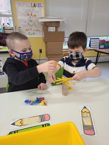 Two kindergarten boys, one wearing a black and white striped shirt and a checked mask, the other wearing a black sweatshirt and printed mask, attempt to balance magnetized letters on a cardboard tube with popsicle sticks on top, looking like a tree.
