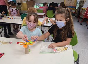 Two kindergarten girls, one with long brown hair, wearing a blue mask and black shirt, and the other wearing a prnted mask and light blue shirt with her hair in a ponytail, balance several letters on the limbs of their little tree made of popsicle sticks on a cardboard tube.