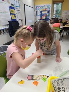 Two kindergarten girls are at a table aancing magnetic letters on their little tree made of a cardboard tube and popsicle sticks on top. One girl with long brown hair is standing and looking down from the top while the other girl with blonde hair in a ponytail puts letters on the tree.