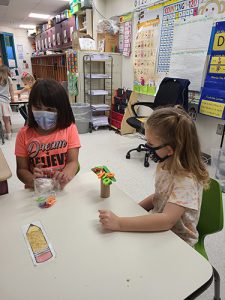 Two girl sitting at a table look at their tree, made of cardboard and popsicle sticks, with several letters on it. One girl has shoulderlength dark hair and is wearing a blue mask and orange shirt. The other girl has long blonde hair and is wearing a flowered shirt and dark mask.