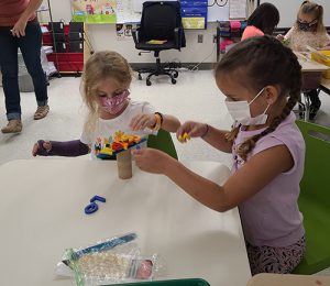 Two girls sit at a table in their kindergarten class. One girl has long dark braids and is wearing a light purple shirt and blue mask. The other is wearing a white shirt and mask and has blonde air. They are putting plastic letters on top of a tree they made of cardboard and popsicle sticks.