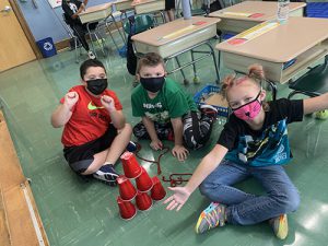 Three students sit on the floor with their six red cups in a pyramid. One of the students on the right holds her arms open wide. All are wearing masks.