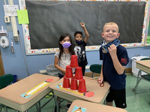 Three students stand around a dsk with six red cups stacked in a pyramid. One boy pulled his mask down to smile. A girl and another boy stand next to the desk, the boy with his arm up in victory.