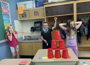 Two third-grade students hold their hands up in victory while one girl off to the left has her hands over her mouth in surprise as their six red cups stand in a pyramid on the desk.