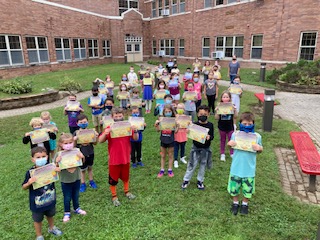 A large group of elementary school children - more than 40, stand in a courtyard holding up certificates. They are all wearing masks. Behind them is a brick building.