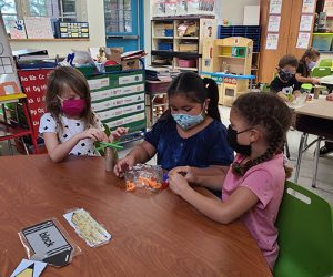 Three kindergarten girls, all with longer dark hair, sit around a table putting magnetic letters onto a cardboard tube. The girls all are wearing masks and are in a classroom.