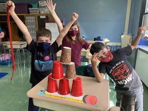 Three third-grade students hold their arms up victorious as their  six cups are stacked in a pyramid on a desk.