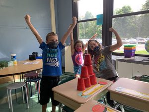 Three third-grade students hold their arms up in victory as they stand by their pyramid of red cups stacked on a desk.