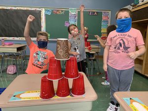 Three students raise their hands in victory showing their six plastic cups stacked on the desk. They are all wearing masks.